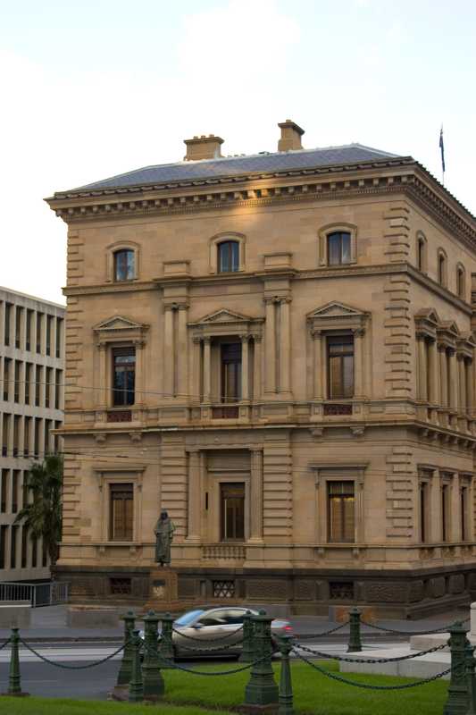 A sandstone building with decorated windows and a dark lower level stands beside a street, with a small statue, passing car and chain fence in front.