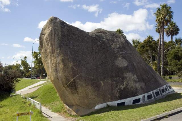 A granite outcrop shaped like a raised dog’s head stands beside a road, with black rectangles along a white band near its base.