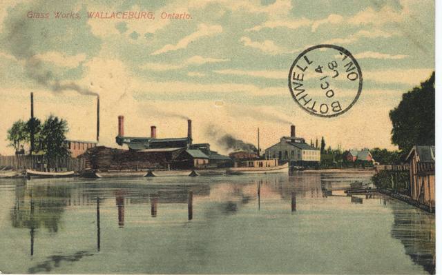 Coloured historical postcard showing glass-factory buildings and chimneys beside the river, with a circular postal mark.
