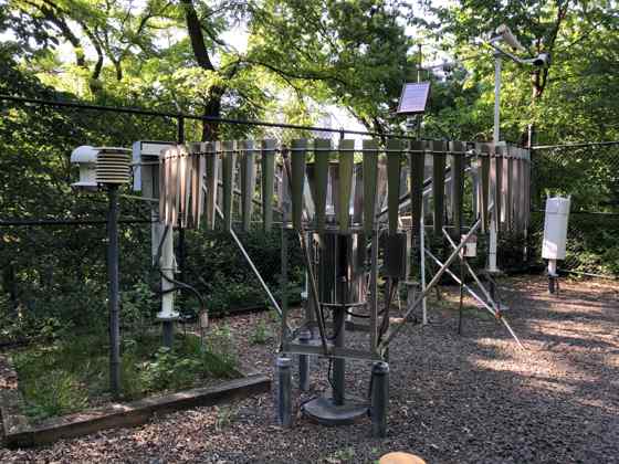 Weather instruments and a circular array of metal slats stand inside a fenced, tree-filled enclosure, with a small solar panel above.