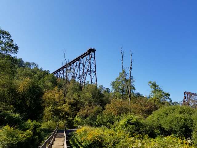 A high steel trestle ends above a wooded valley, with a small wooden footbridge in the foreground.