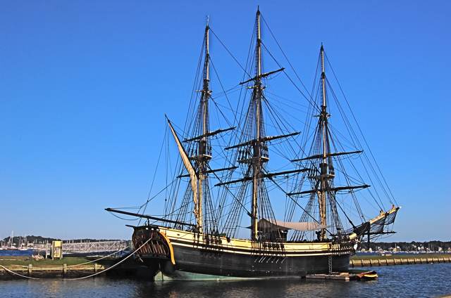 A black-and-yellow ship has three tall masts with dense rigging, moored alongside a wharf in blue water.