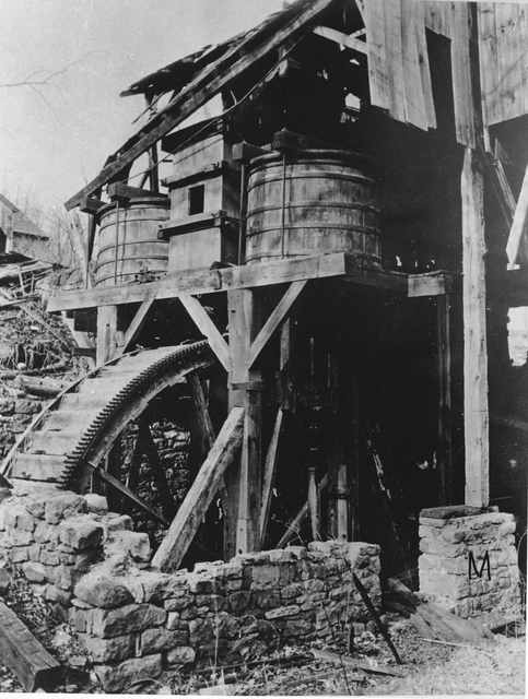 Black-and-white historical view of an incomplete wheel below two wooden tubs, with broken roofing and exposed supporting timbers.