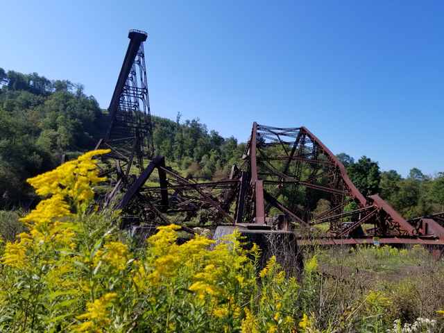 Fallen steel frameworks lie amid vegetation below the surviving end of a tall trestle.