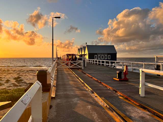 Rails and a red train beside a dark visitor building at the shore end of Busselton Jetty, with the jetty extending into the sea.